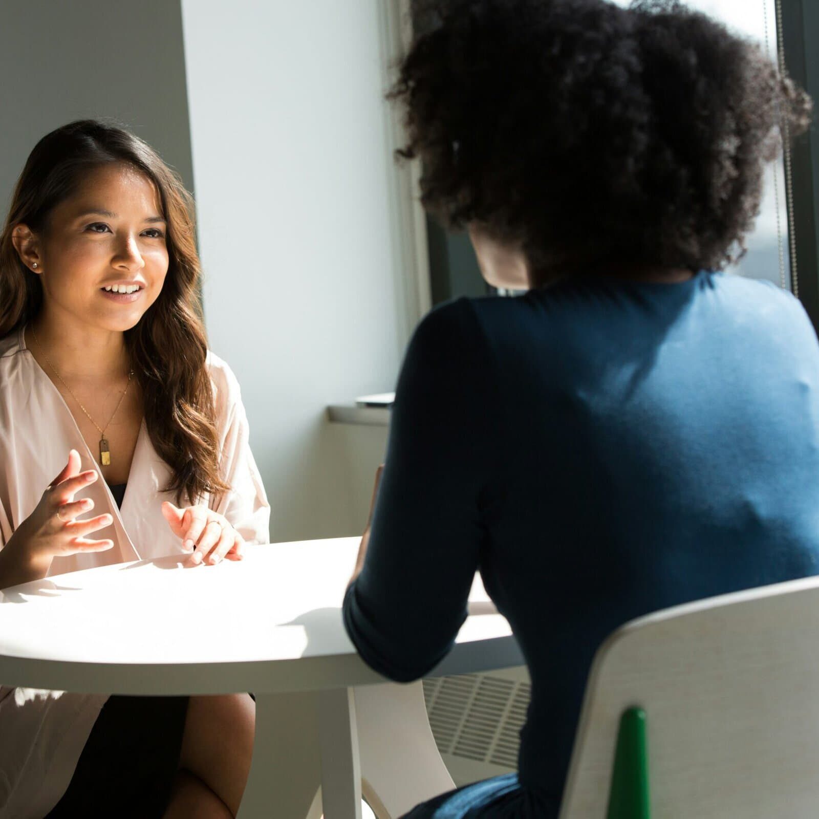 Two woman sit at a table in a semi professional setting talking to each other.