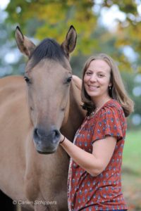 Katie Navarra Bradley ACC headshot photo, smiling woman with light brown hair wearing red top standing next to fawn colored horse with dark hair outside