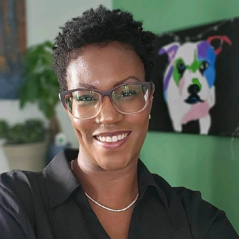 Ayana King headshot photo, smiling woman wearing glasses and black shirt in front of green wall and painting of a dog