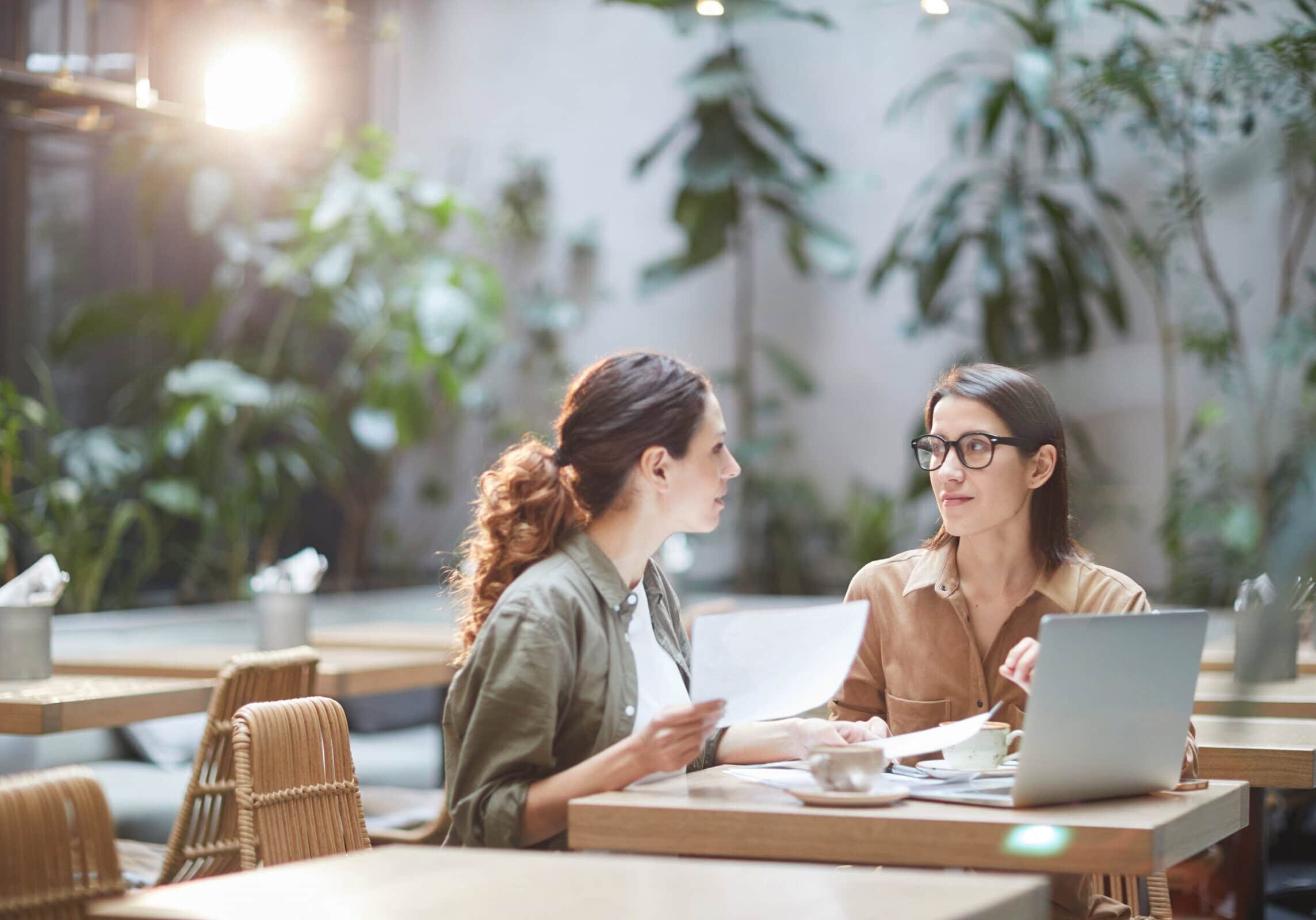 An experienced coach in glasses is meeting with an aspiring coach in an empty sunlit cafe with plants, reviewing coaching materials, and using a laptop.