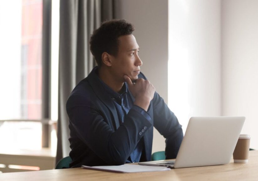Young professional contemplating career growth while seated at a table with a laptop, symbolizing the potential benefits of coaching for career advancement.