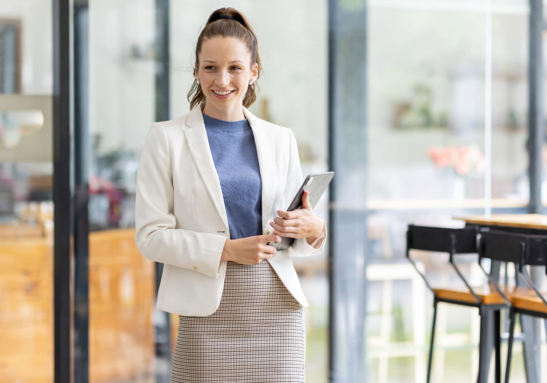 A smiling professional businesswoman is standing in an office with a tablet.