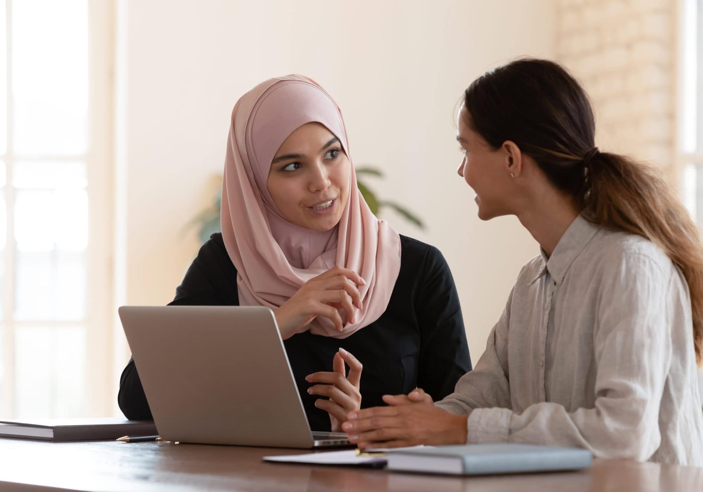 Two women having a focused discussion at a table with a laptop open in front of them.