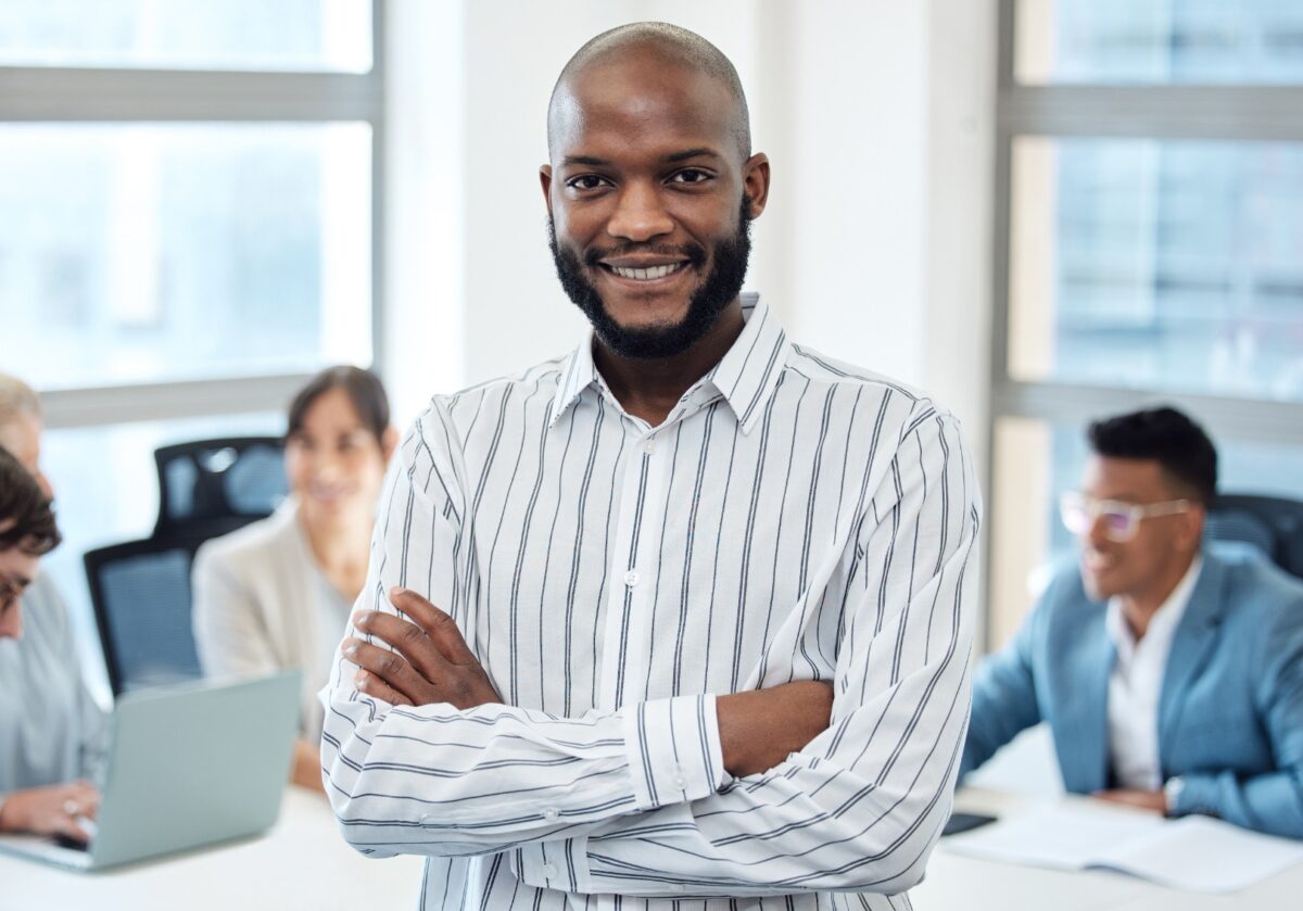 Portrait of a young, African American businessman standing with his arms crossed in an office with his colleagues in the background sitting around a table.