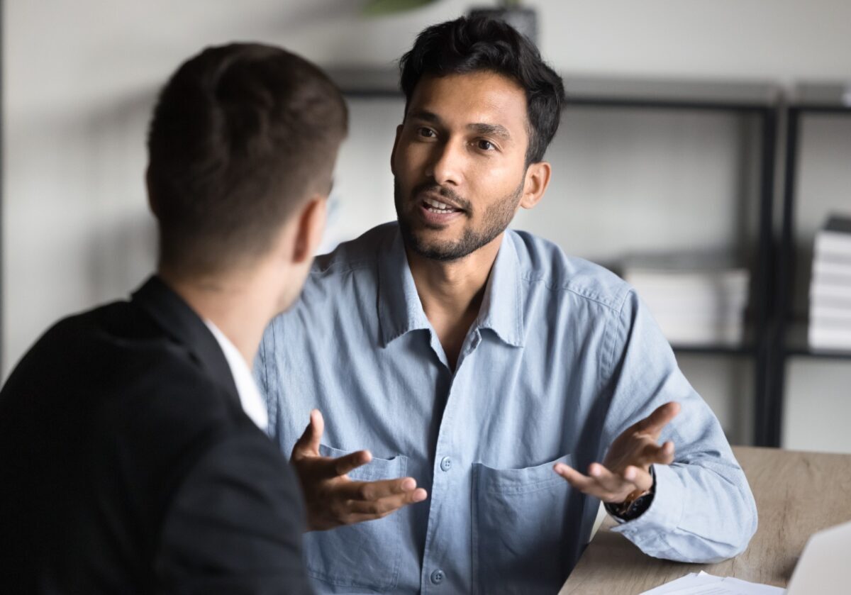 Two men are sitting at a table having a serious conversation; one man is speaking with expressive hand gestures while the other listens attentively.