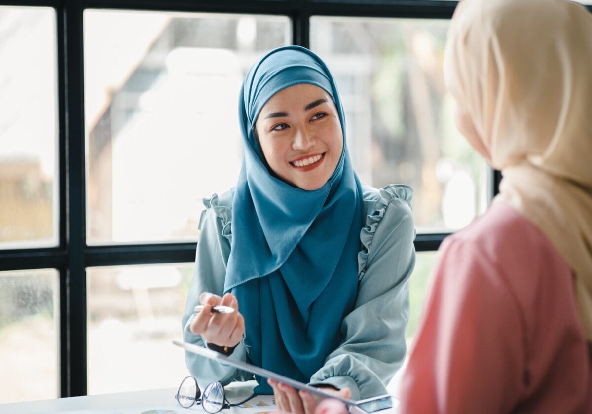 Two women wearing hijabs discuss work while looking at a tablet in a bright office.