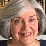 Judy Feld headshot photo, woman with gray hair smiling in front of bookshelf
