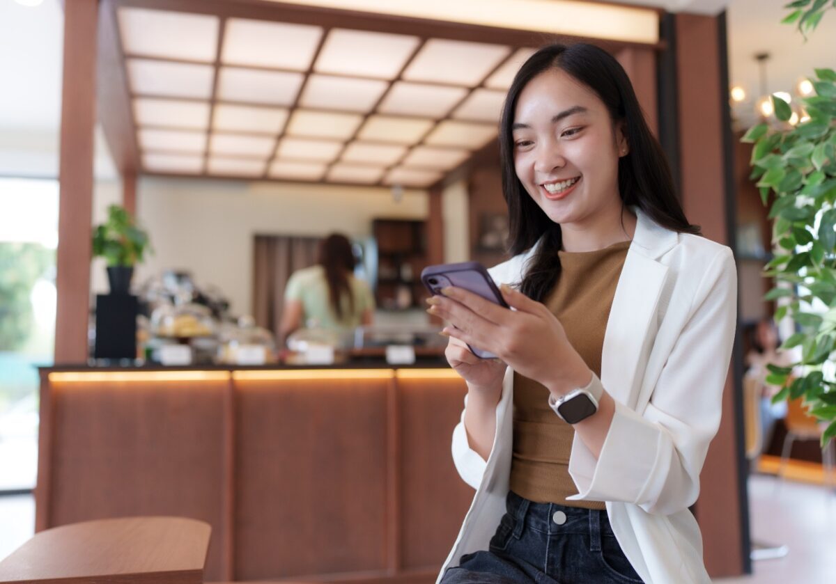 Young Asian businesswoman is sitting in a cafe, smiling while using her smartphone, enjoying her break time in a modern and stylish setting.