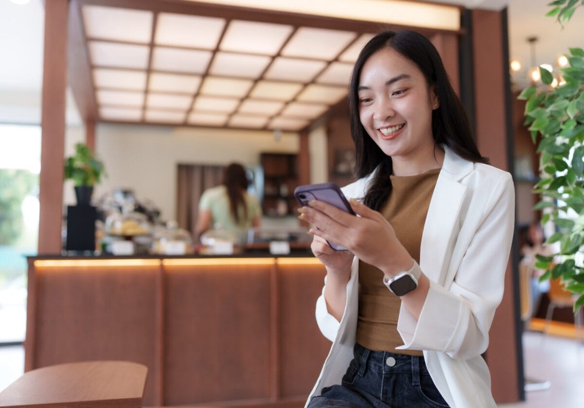 Young Asian businesswoman is sitting in a cafe, smiling while using her smartphone, enjoying her break time in a modern and stylish setting.