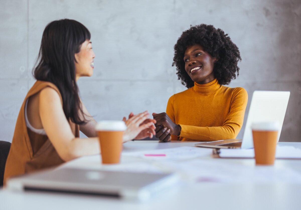 Two diverse women smile and talk across a table in an office, surrounded by papers and laptops.