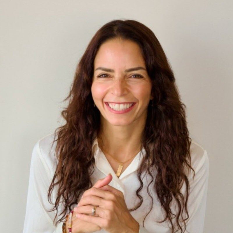 Mona Seleim ACC headshot photo, smiling woman with long brown hair wearing a white button up shirt with hands clasped in front of white background