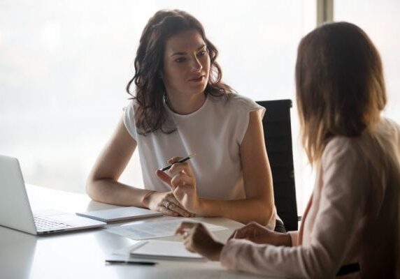 coach listening to a client speak while sitting at a table with a laptop and notepads