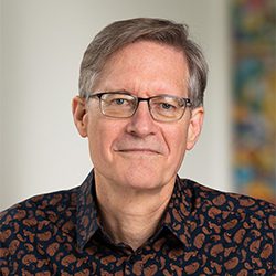 Paul Boehnke PCC ELI MP headshot photo, gray haired man with glasses and dark collared shirt smiling, blurred background