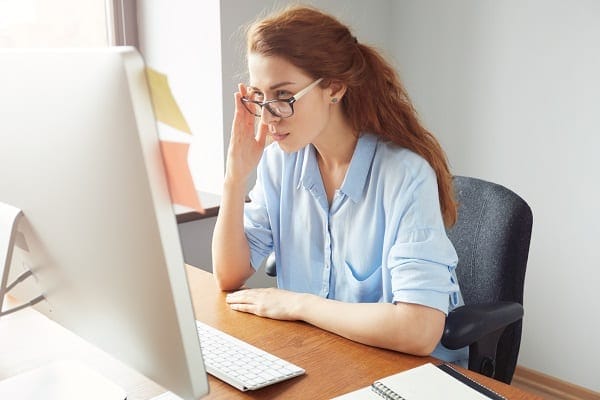 woman with glasses on sitting at a desk looking at a website on a computer monitor