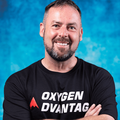 Patrick McKwoen headshot photo, smiling man wearing black long sleeve shirt with logo Oxygen Advantage in front of blue background